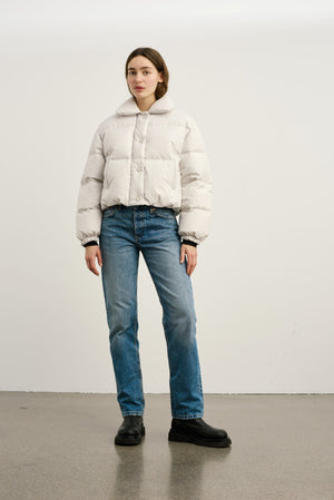 A model wears a light beige puffer jacket paired with blue jeans and black shoes, showcasing a stylish winter look by MEOTINE.