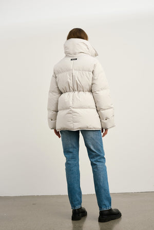A model wearing a light beige puffer jacket from MEOTINE, paired with blue jeans, standing against a plain background.