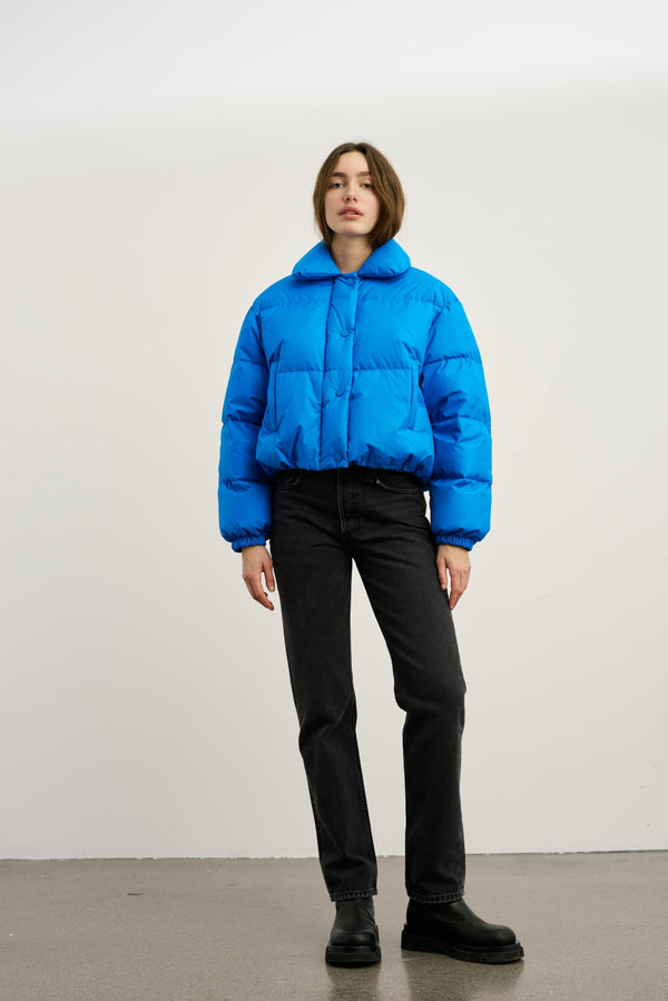A model wears a vibrant blue puffer jacket from MEOTINE, paired with black jeans and chunky boots, set against a neutral background.