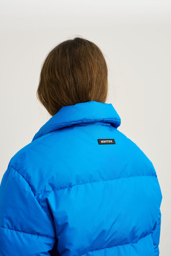 A woman with long hair wears a vibrant blue puffer jacket featuring the MEOTINE logo on the back, showcasing its stylish design.