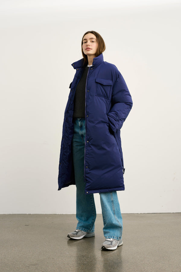 A model showcases a long navy blue puffer coat with a high collar and front pockets, paired with relaxed denim jeans and sneakers. MEOTINE.