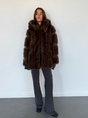 A model wearing a luxurious brown fur coat by MEOTINE, paired with gray flared pants, standing against a plain background.