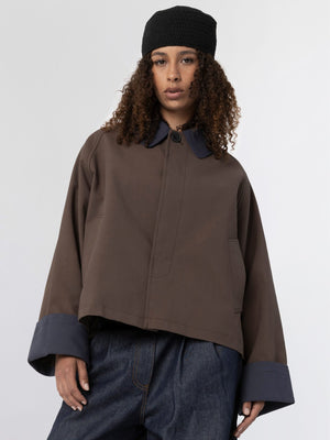 A model wears a brown MEOTINE jacket with a navy collar and cuffs, paired with dark denim pants and a black beanie.