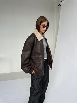 A model wears a brown leather jacket with a fur collar, paired with gray pants and sunglasses, showcasing MEOTINE's stylish design.
