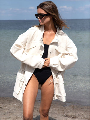 A model wearing a cream oversized jacket over a black swimsuit, standing on a beach with calm water in the background. MEOTINE.