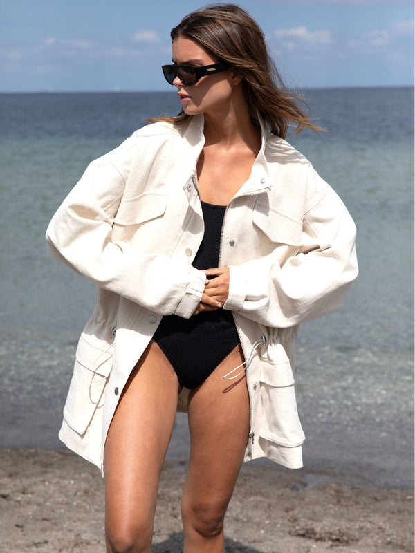 A model wearing a cream oversized jacket over a black swimsuit, standing on a beach with calm water in the background. MEOTINE.