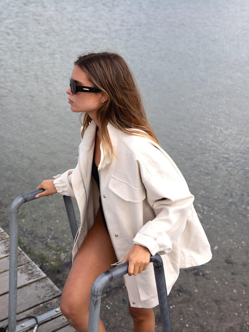 A model wearing a cream oversized jacket over a black swimsuit, standing on a beach with calm water in the background. MEOTINE.