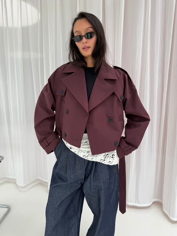 Chic model wearing a burgundy cropped trench coat over a white lace top, paired with wide-leg denim pants and sunglasses. MEOTINE.