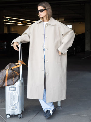 A model in a beige oversized coat stands beside a silver suitcase, exuding a chic, modern style with sunglasses and casual jeans. MEOTINE.