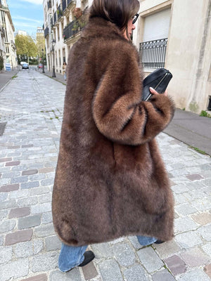 A woman walks down a cobblestone street wearing a luxurious brown MEOTINE fur coat, paired with casual jeans and a sleek black bag.
