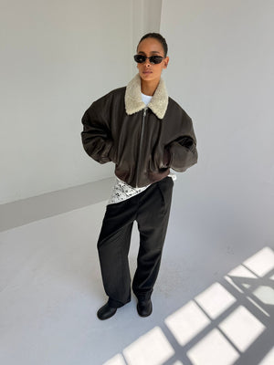 A model showcases a stylish brown bomber jacket with a faux fur collar, paired with black trousers and sunglasses, representing MEOTINE.