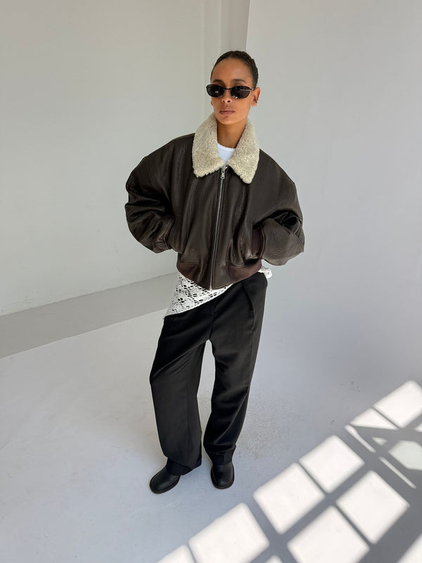 A model showcases a stylish brown bomber jacket with a faux fur collar, paired with black trousers and sunglasses, representing MEOTINE.