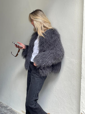 A woman in a fluffy gray MEOTINE jacket checks her phone, styled with a casual white top and black pants against a light wall.