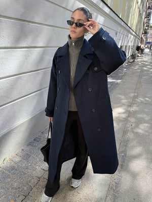 A person wearing a navy oversized coat, gray sweater, and black pants, accessorized with sunglasses and a black bag, showcasing MEOTINE style.