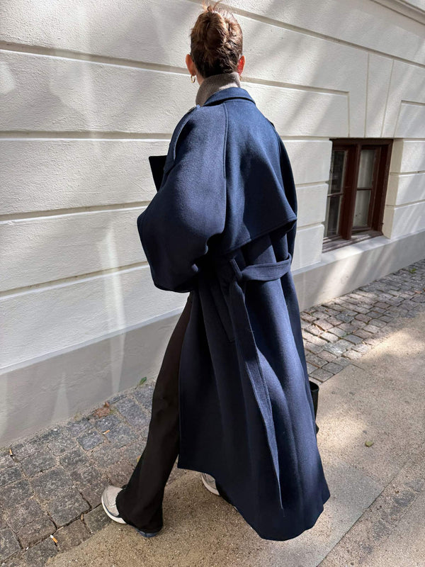 A woman walks down a cobblestone path wearing a stylish navy coat with a belt, showcasing MEOTINE's elegant design.