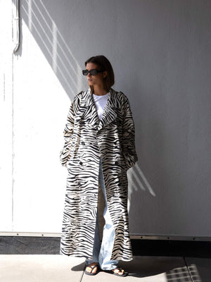 A model wearing a stylish zebra print coat by MEOTINE, paired with light jeans and sunglasses, stands against a white wall.