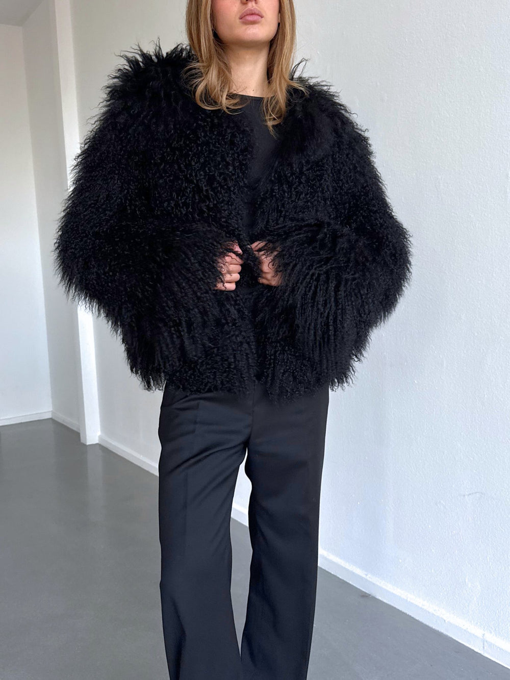 A model showcases a luxurious black fluffy coat by MEOTINE, paired with sleek black pants, set against a minimalist backdrop.