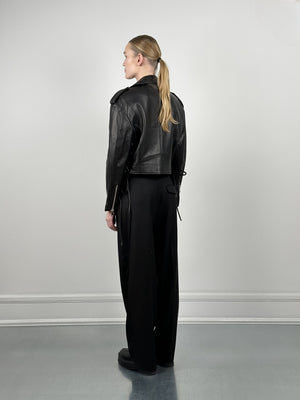 A woman in a cropped black leather jacket and loose black pants stands with her back to the camera, showcasing MEOTINE's stylish design.