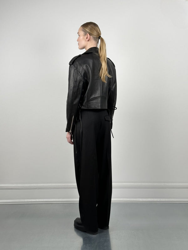 A woman in a cropped black leather jacket and loose black pants stands with her back to the camera, showcasing MEOTINE's stylish design.