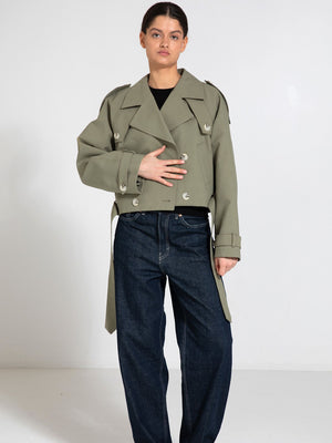 A model wears a stylish cropped olive green trench coat with oversized buttons, paired with loose-fitting dark jeans. MEOTINE.