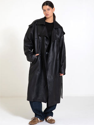 A model showcases a stylish black leather trench coat by MEOTINE, paired with casual jeans and cozy footwear.