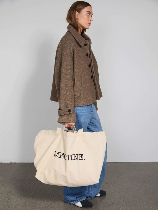 A model wearing a brown textured jacket and blue jeans carries a large beige tote bag with "MEOTINE." printed on it.