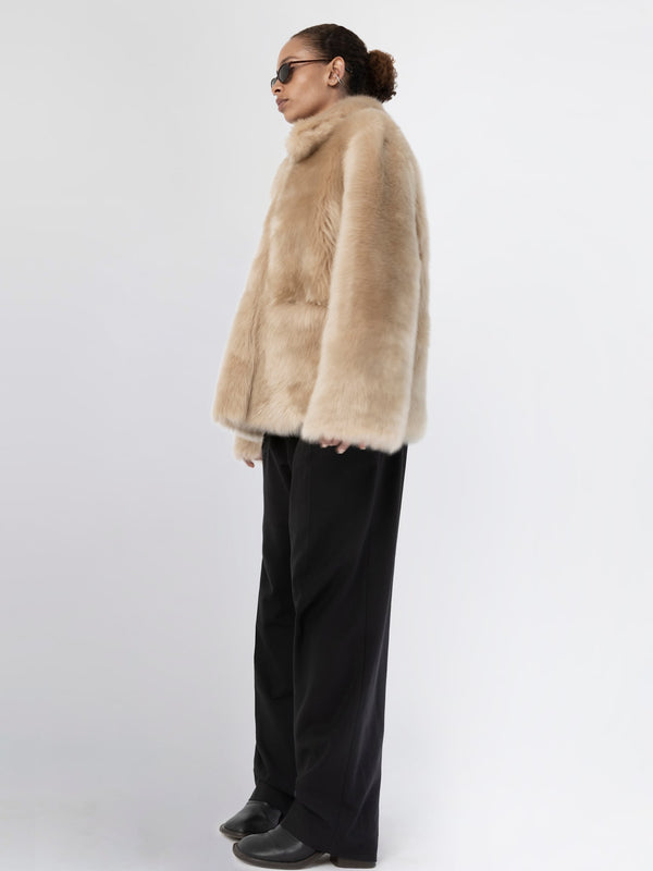 A model showcases a luxurious beige faux fur coat by MEOTINE, paired with sleek black trousers and stylish sunglasses.