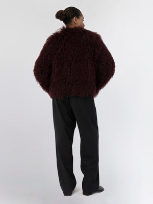 A model showcases a cozy, burgundy MEOTINE faux fur jacket paired with black trousers, highlighting a stylish, relaxed look.