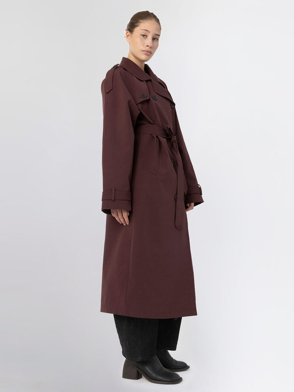 A model showcases a long burgundy trench coat with a belt, featuring a classic collar and button details, by MEOTINE.