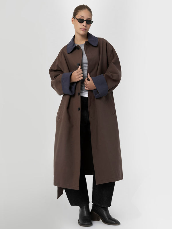 A model wears a stylish oversized brown coat with navy collar and cuffs, paired with a gray top and black pants. MEOTINE.