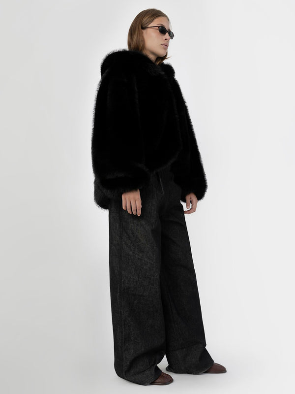 A model wearing a black faux fur coat with oversized sleeves, paired with wide-leg black trousers and stylish sunglasses by MEOTINE.