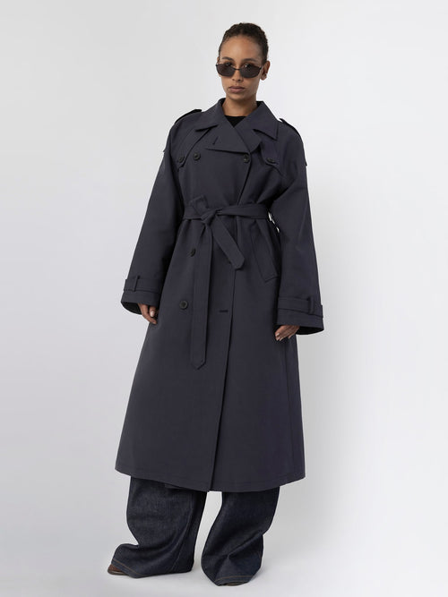 A model showcases a stylish dark trench coat with a belt, paired with wide-leg jeans and sunglasses, representing MEOTINE's chic aesthetic.