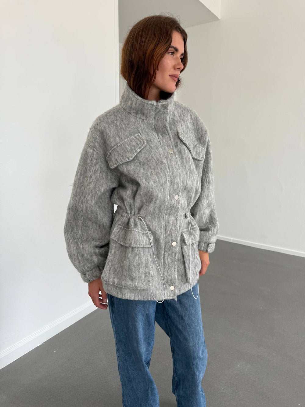 A model showcases a stylish gray MEOTINE jacket with a high collar, front pockets, and a cinched waist, paired with blue jeans.
