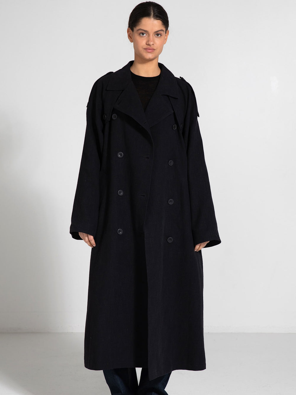 A model showcases a long, dark coat with a relaxed fit and oversized sleeves, styled with jeans and sleek shoes, by MEOTINE.