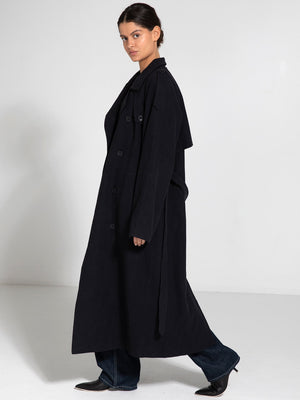 A model showcases a long, dark coat with a relaxed fit and oversized sleeves, styled with jeans and sleek shoes, by MEOTINE.