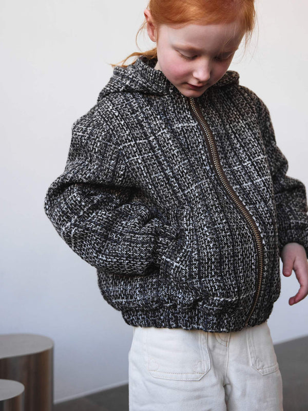 A young girl in a cozy, textured black and white zip-up jacket with a hood, paired with light-colored pants. MEOTINE brand style.