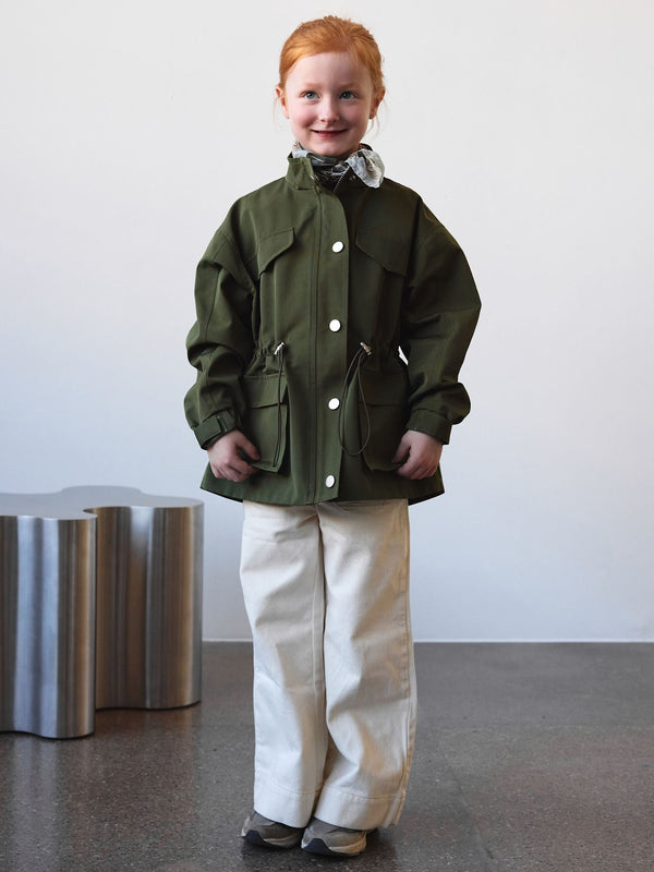 A child wearing a green MEOTINE jacket with pockets, paired with white pants, stands against a minimalist background.