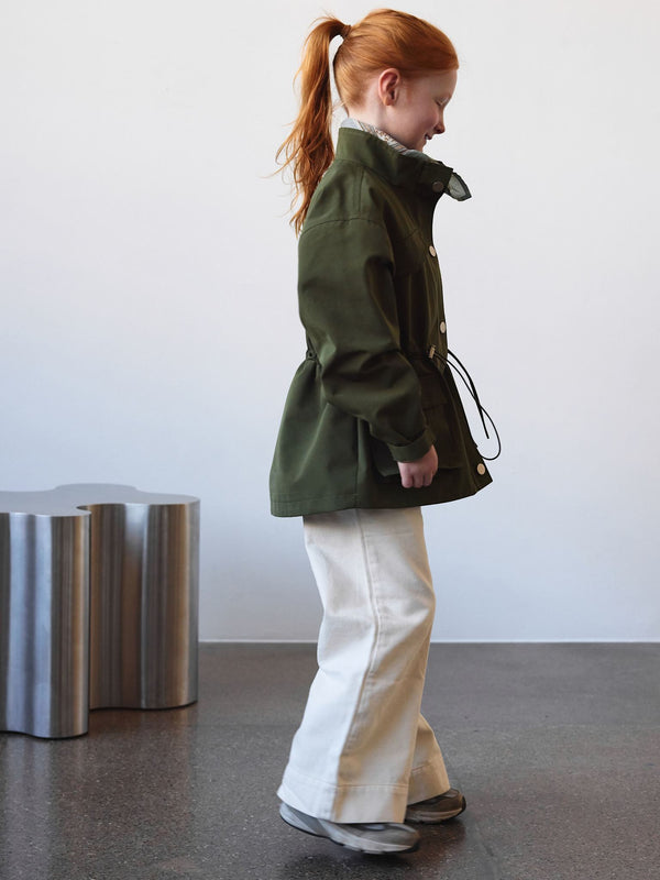 A girl in a green MEOTINE jacket and cream pants stands against a minimalist backdrop, showcasing a stylish, casual look.