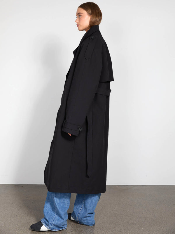 A model showcases a stylish black trench coat from MEOTINE, paired with wide-leg jeans, set against a minimalist backdrop.