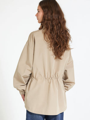 A woman with long, wavy hair wears a beige MEOTINE jacket, featuring a cinched waist and relaxed fit, viewed from the back.
