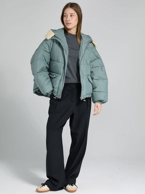 A model wears a cozy, oversized teal puffer jacket with a fur-lined hood, paired with black wide-leg pants. MEOTINE style.