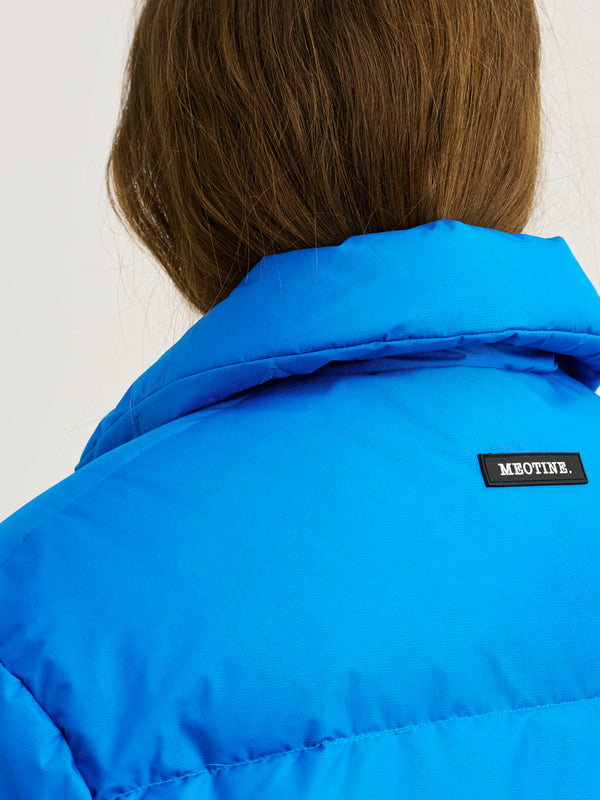 A close-up of a woman's back wearing a vibrant blue jacket with the MEOTINE logo on the collar, showcasing its stylish design.