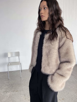 A model showcases a soft, beige faux fur jacket by MEOTINE, styled with a black top and relaxed trousers in a minimalist setting.