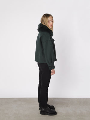 A model stands sideways wearing a dark green MEOTINE jacket with a fur collar, paired with black jeans and chunky boots.