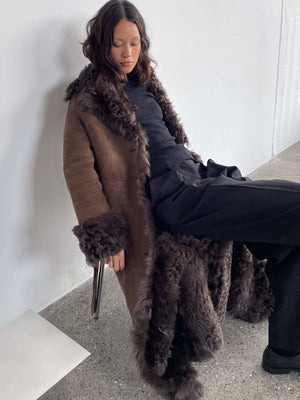 A model wearing a luxurious brown fur coat with a plush collar and cuffs, styled with black pants, showcasing MEOTINE's elegance.