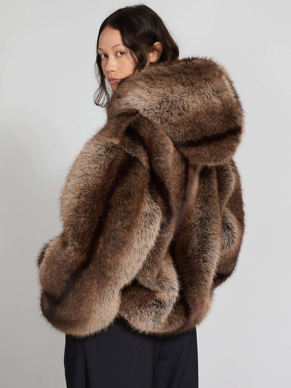 Cozy brown faux fur jacket with a hood, paired with black trousers, worn by a model sitting on a chair. MEOTINE style.