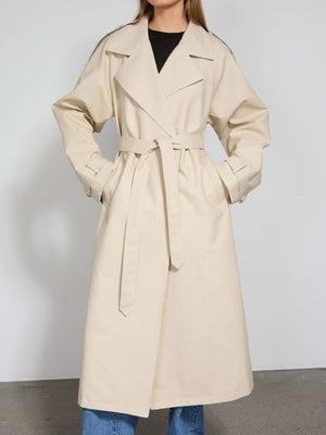 A stylish beige trench coat with a belted waist, perfect for layering, showcased by a model against a neutral background. MEOTINE.