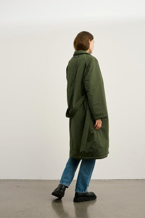 A model wearing a long, olive green MEOTINE coat, paired with blue jeans and black boots, stands against a plain background.
