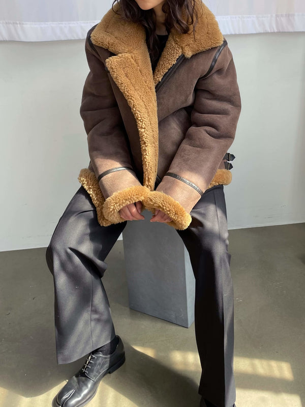 A model sits wearing a stylish brown shearling jacket with a cozy tan collar, paired with black trousers and boots. MEOTINE.