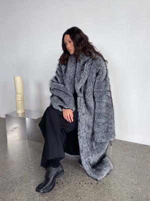 A model sits elegantly in a plush gray MEOTINE fur coat, paired with black attire, showcasing a chic winter look.